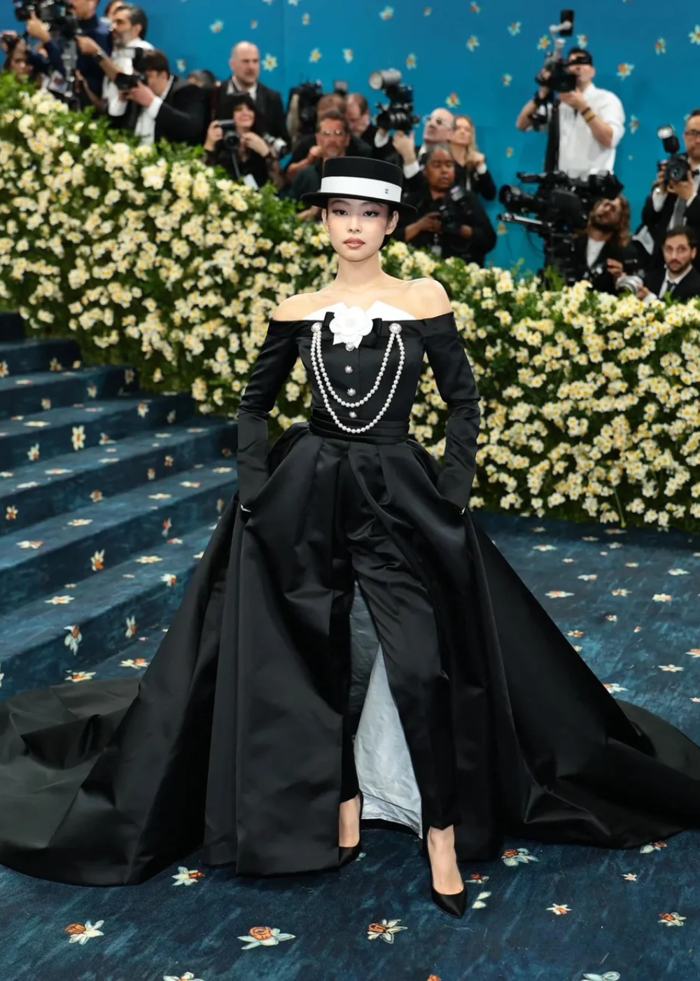 Jennie vestida de preto com pérolas no pescoço e chapéu elegante, em evento fashion.
