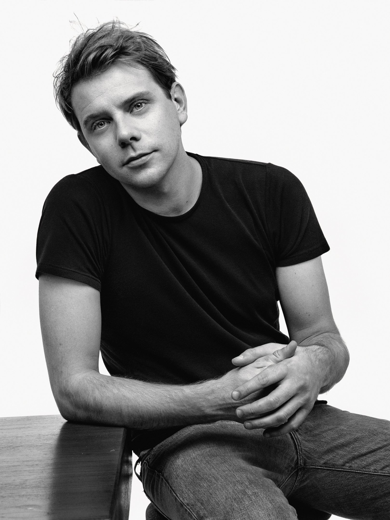 Jonathan Anderson com camiseta preta em pose casual em ambiente minimalista. Moda de Luxo