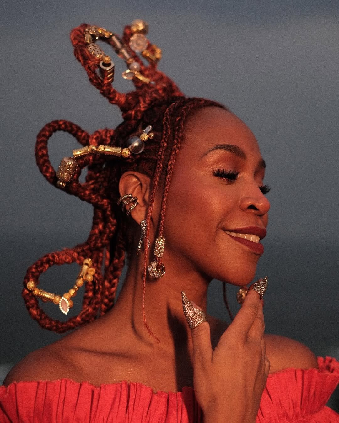Karol Conká com penteado elaborado e tranças decorativas, usando vestido vermelho e sorrindo levemente.