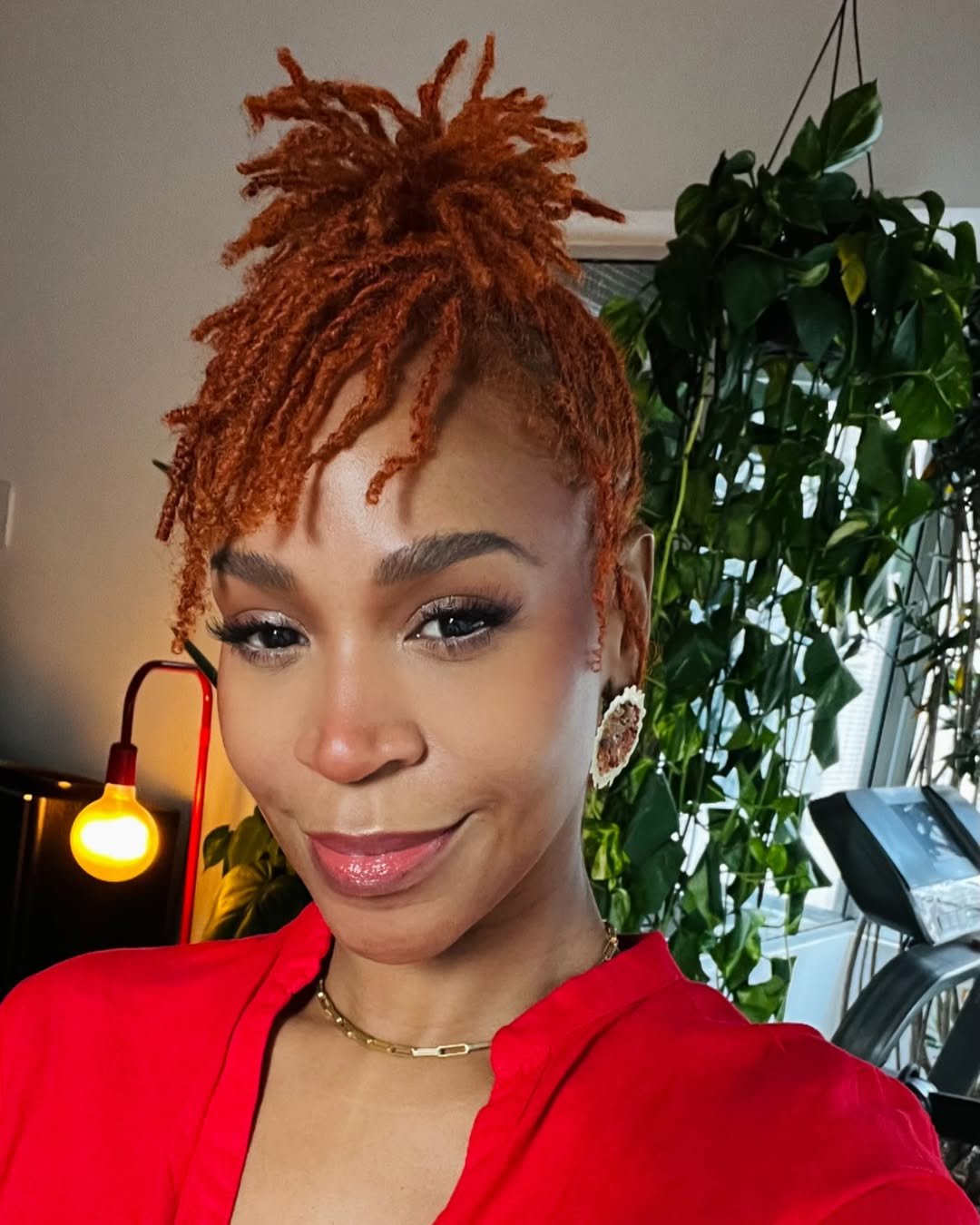 Karol Conká sorrindo com cabelo ruivo e curto, usando camisa vermelha. Ao fundo, há plantas e uma luminária acesa.