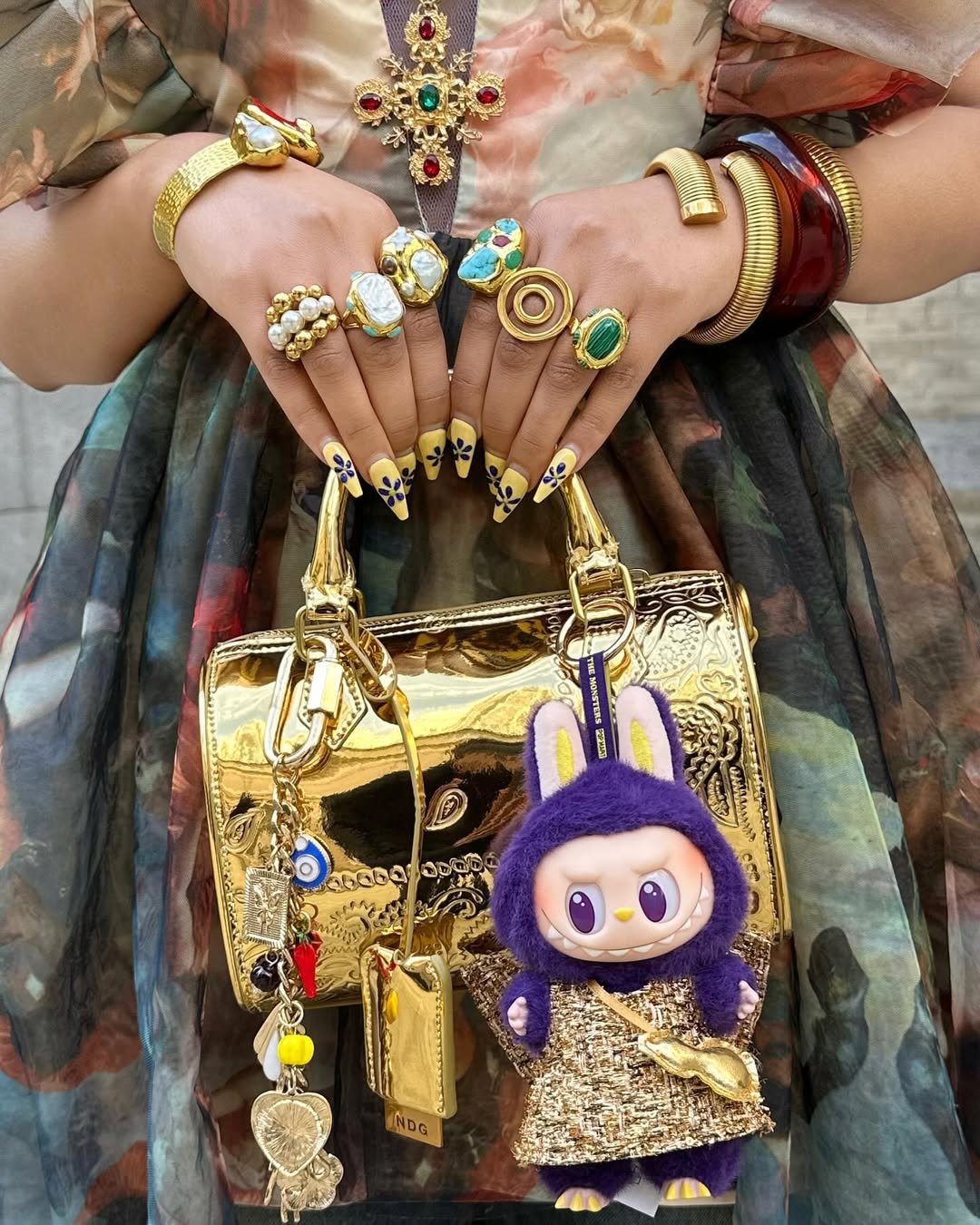 Unhas decoradas com flores e anéis chamativos seguram bolsa dourada. Chaveiro Labubu roxo e dourado pendurado.