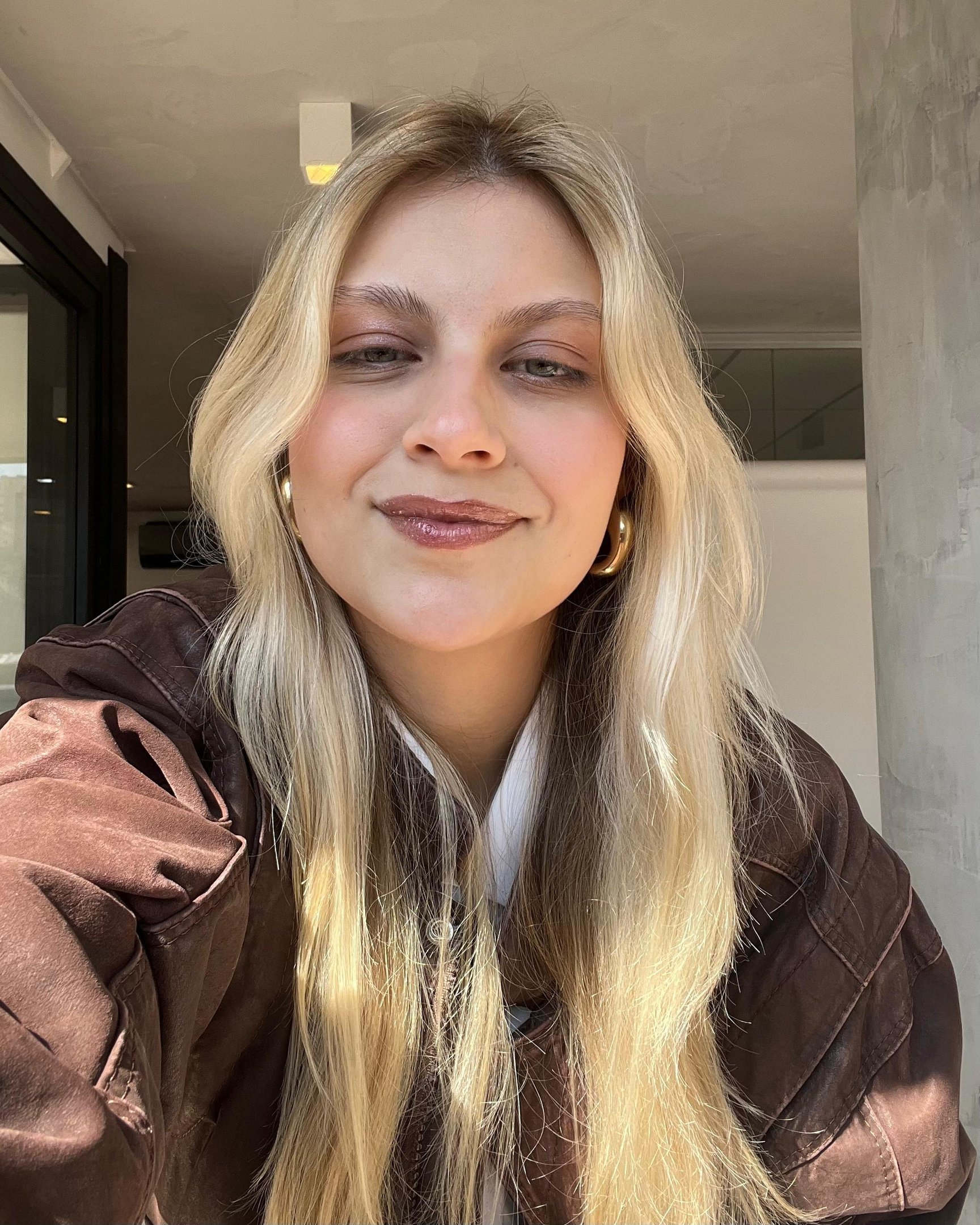 Loira sorrindo suavemente em ambiente interno iluminado, brincos de argola dourados destacam-se.