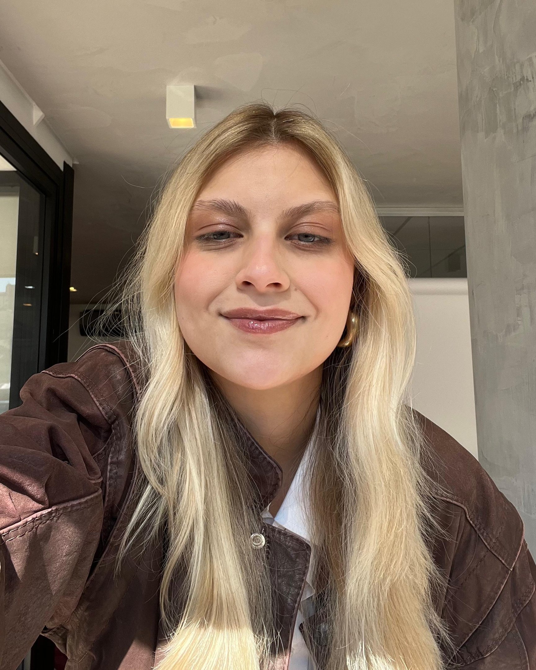 Pessoa loira sorrindo levemente, usando batom brilhante e jaqueta marrom.