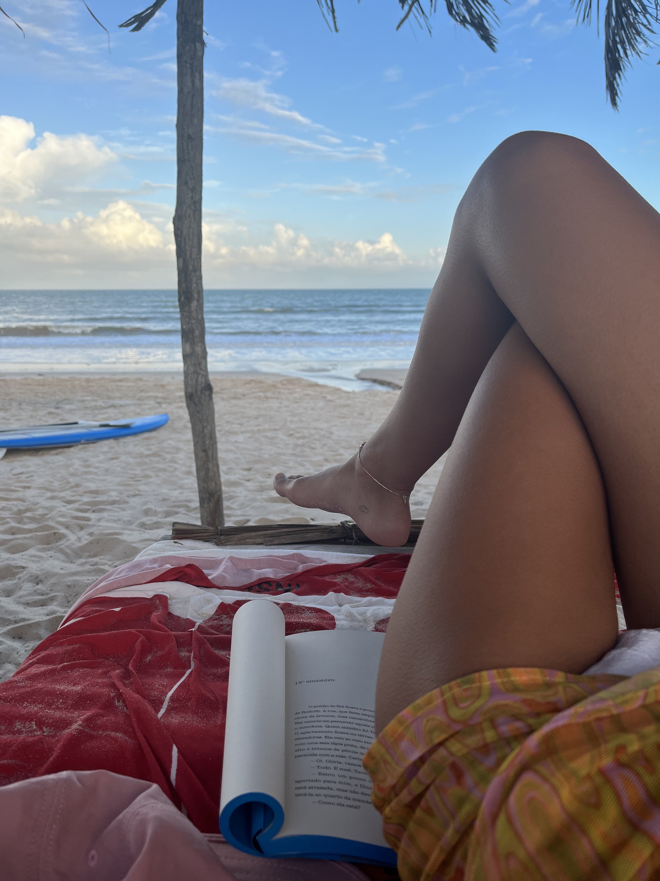 Praia com livro aberto sobre toalha vermelha, pernas cruzadas e vista do mar ao fundo.
