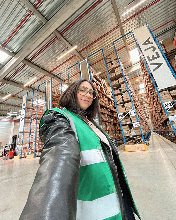Pessoa de colete verde em um armazém com estantes e caixas, sinalização VEJA visível no fundo.