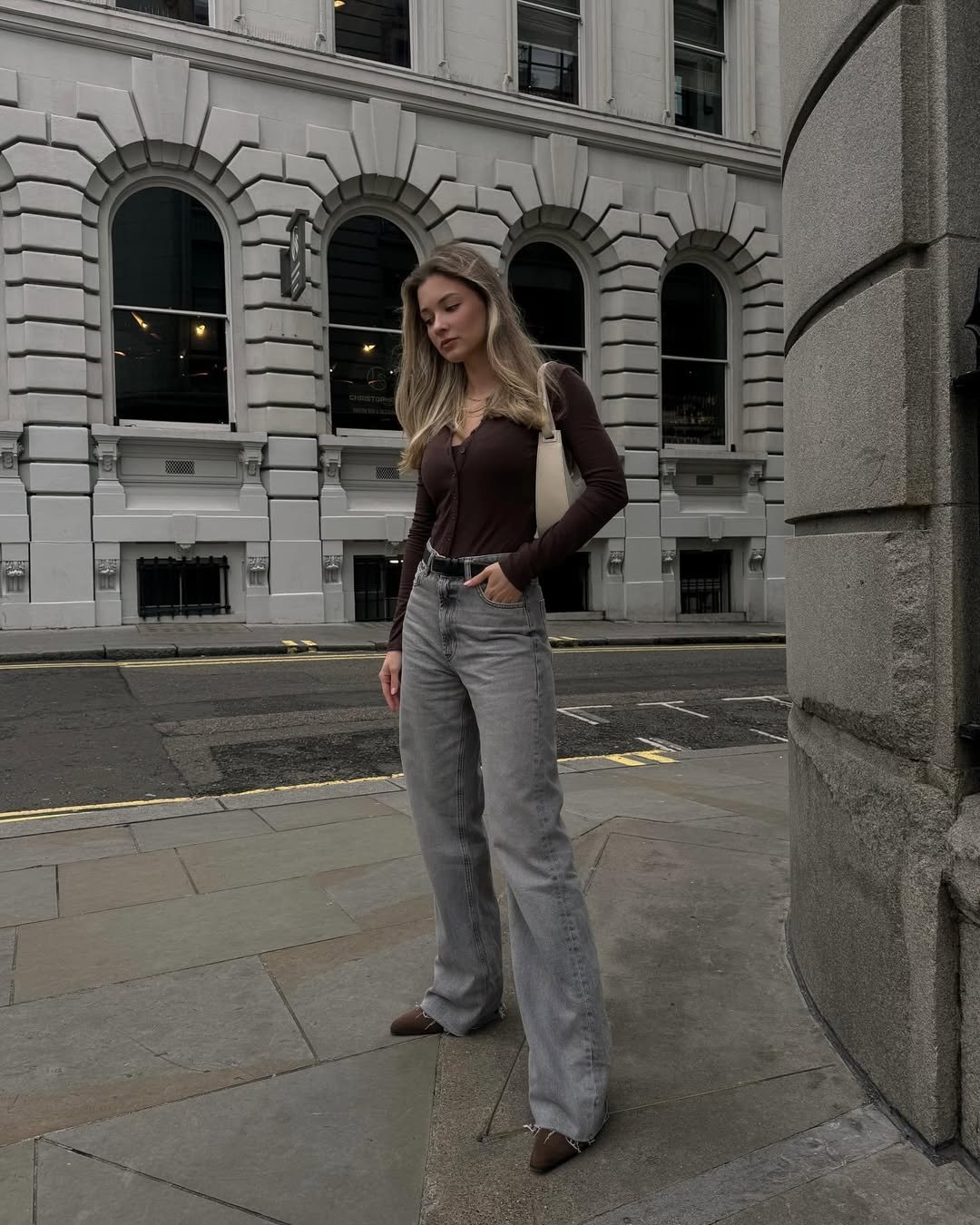 A pessoa está na calçada em frente a um prédio histórico, usando look com bota cano curto, calça jeans cinza e blusa marrom.