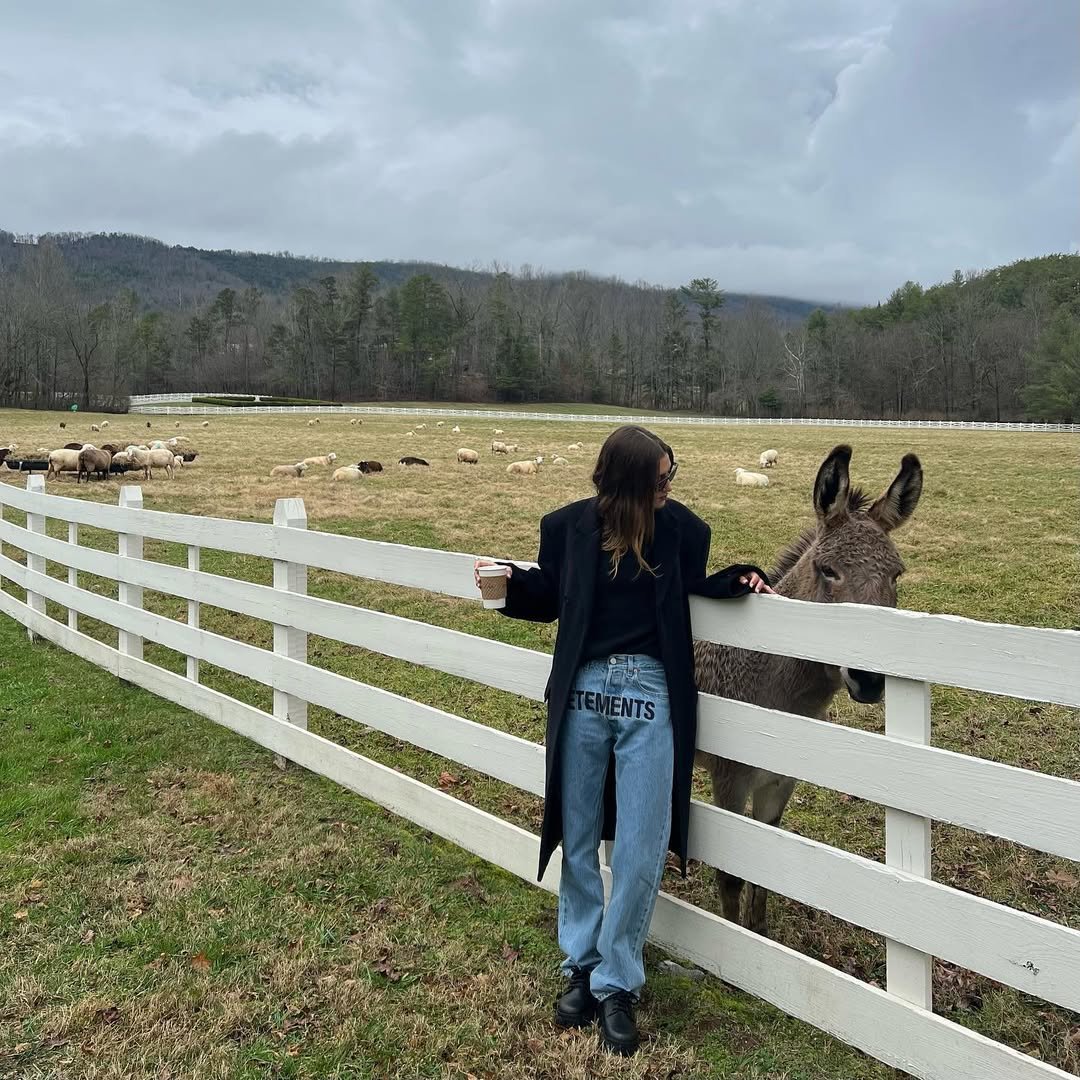 Hailey Bieber usando look com bota cano curto e calça jeans, encostada em cerca branca, segurando copo e ao lado de burro.