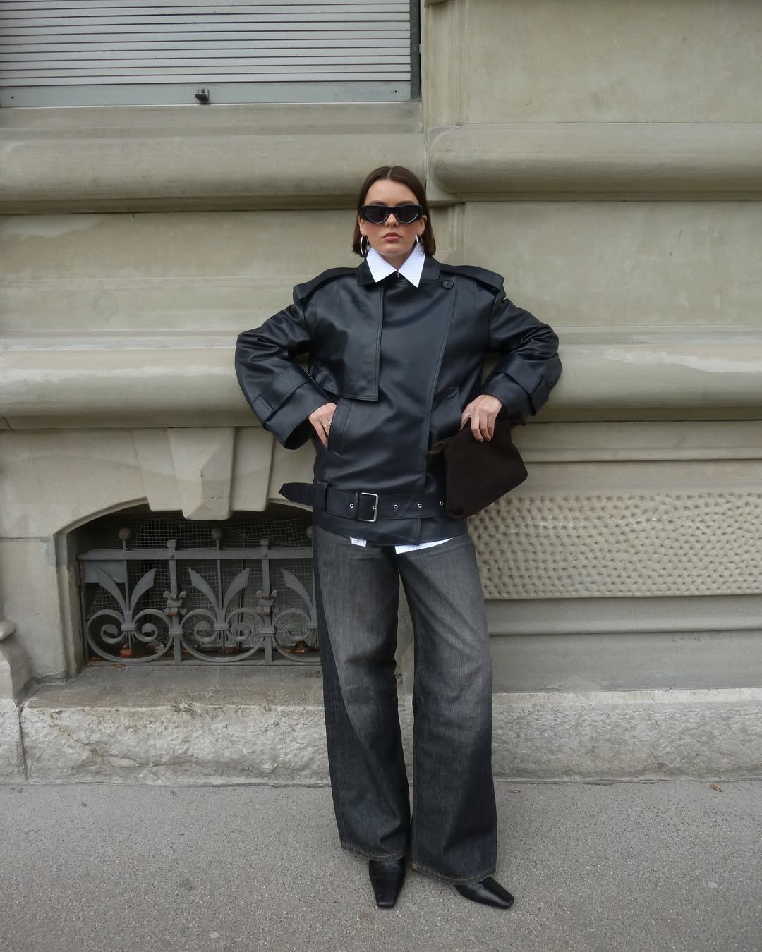 Pessoa usando jaqueta de couro preta, calça jeans larga e botas de bico fino, em frente a um edifício.
