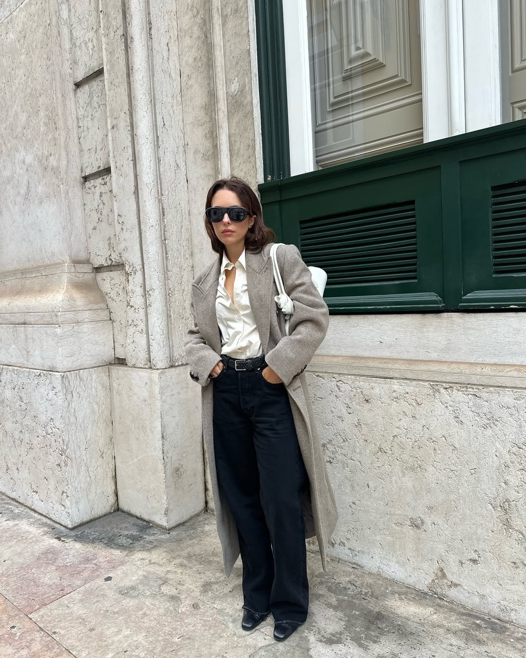 Pessoa de casaco longo bege, camisa clara, calça preta e botas de cano curto em frente a uma fachada branca.
