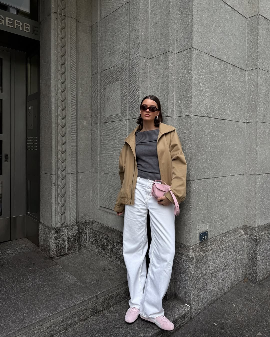 Pessoa com look com calça branca, blusa cinza e jaqueta bege. Ela usa óculos escuros e segura uma bolsa rosa.