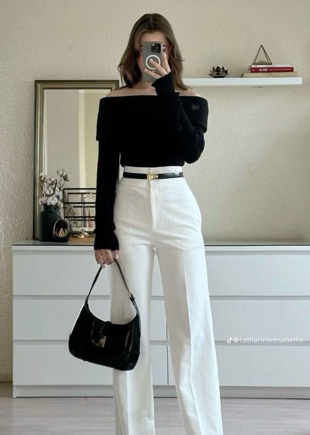 Pessoa fotografando o look com calça branca, blusa preta ombro a ombro, cinto e bolsa pretos em espelho.