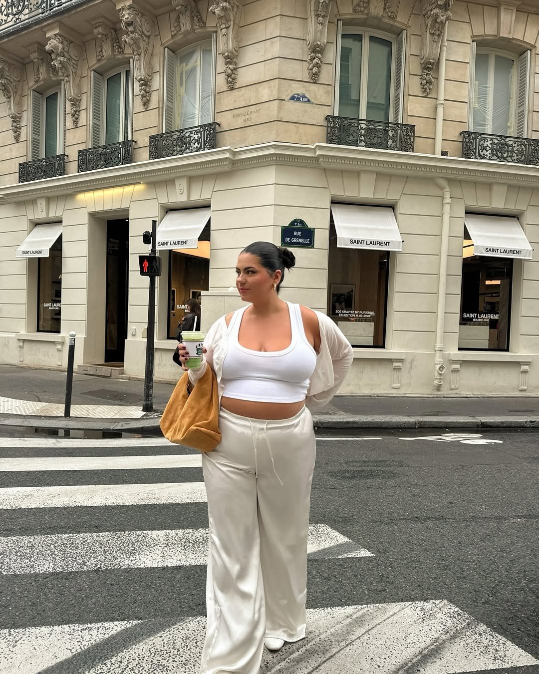 Pessoa com look com calça branca e top, segura bebida em frente a prédio clássico com lojas.