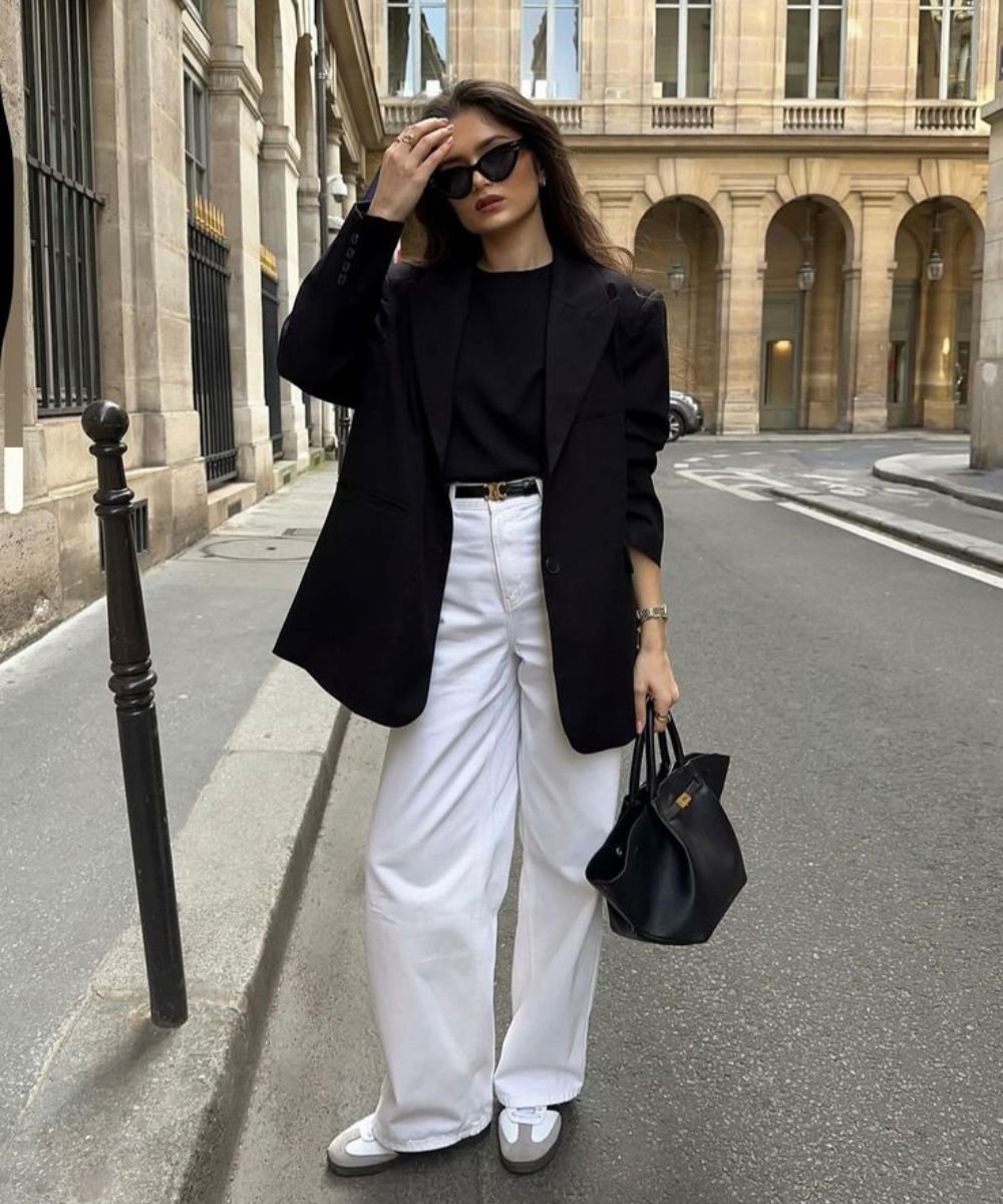 Pessoa usando look com calça branca, blazer preto e camiseta preta, segurando óculos de sol e bolsa preta.