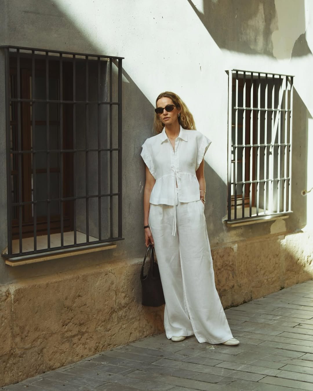 Pessoa com look com calça branca e blusa combinando, encostada em parede iluminada.