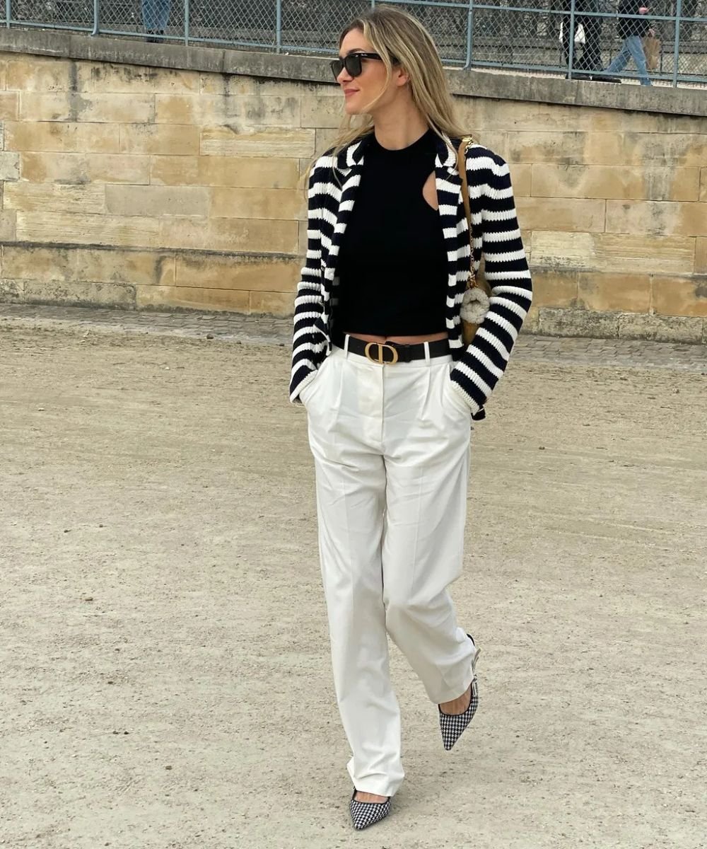 Pessoa com look elegante, usando calça branca, suéter listrado e óculos escuros, caminhando em área externa.