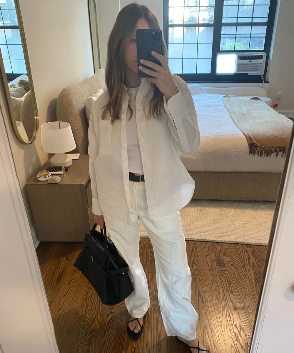 Pessoa em look casual, com camisa e calça branca, segurando bolsa preta em quarto com espelho.
