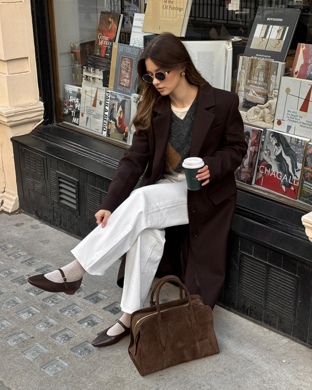 Pessoa usando óculos e segurando bebida, veste um look com calça branca e trench coat na frente de uma vitrine de livros.