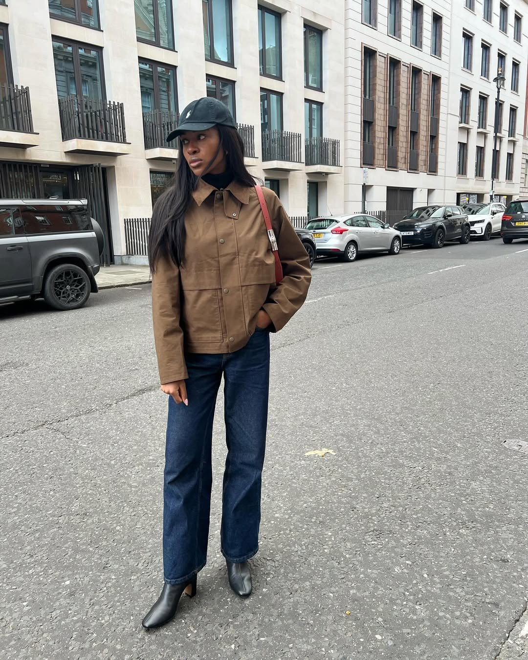 Pessoa usando look com bota cano curto e calça jeans, jaqueta marrom e boné preto em ambiente urbano.