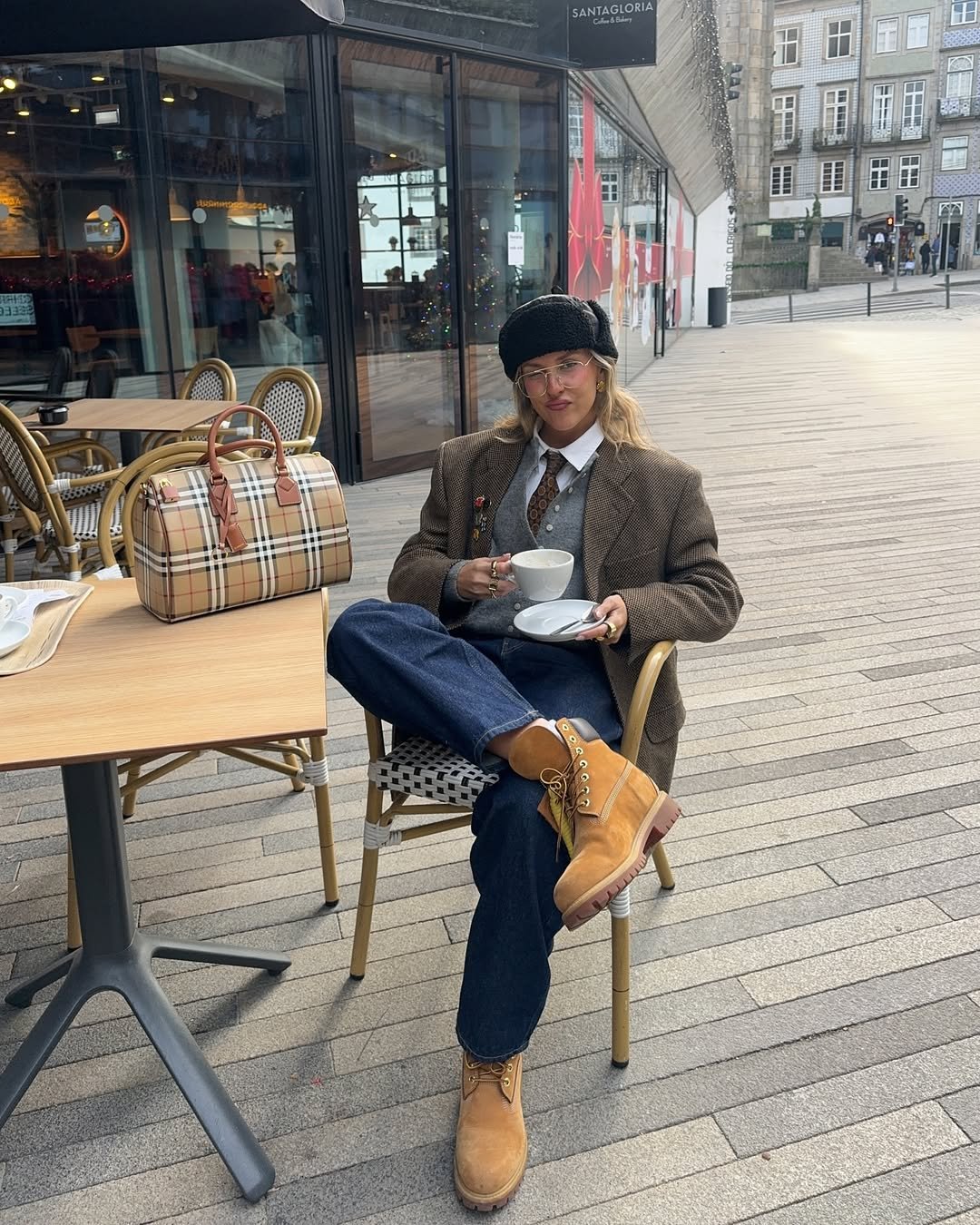Pessoa sentada em cadeira de café, usando look com bota de cano curto e calça jeans, segurando xícara de café. Ao lado, bolsa xadrez.