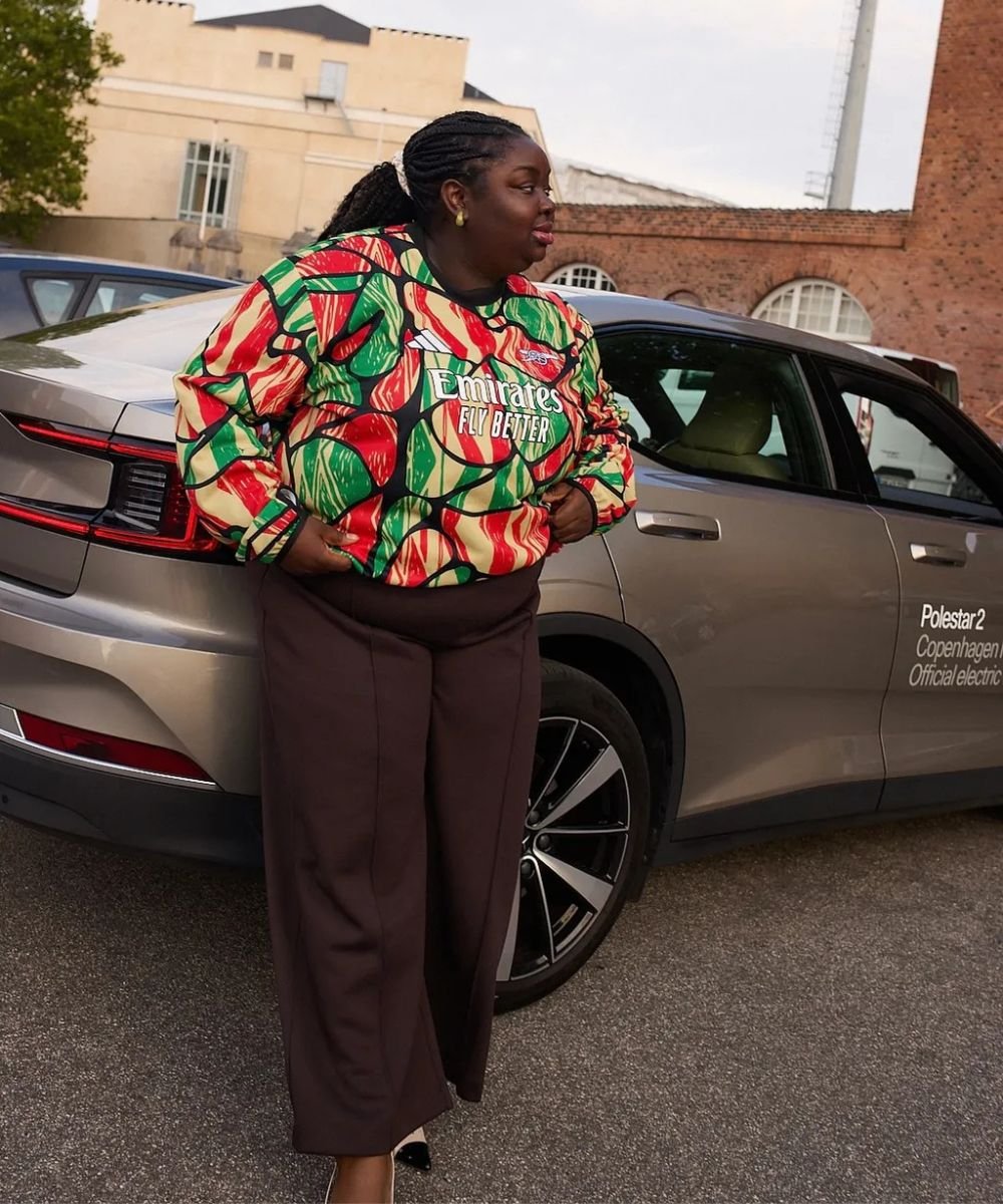 Pessoa com moletom estampado colorido e calça marrom, encostada em um carro cinza.