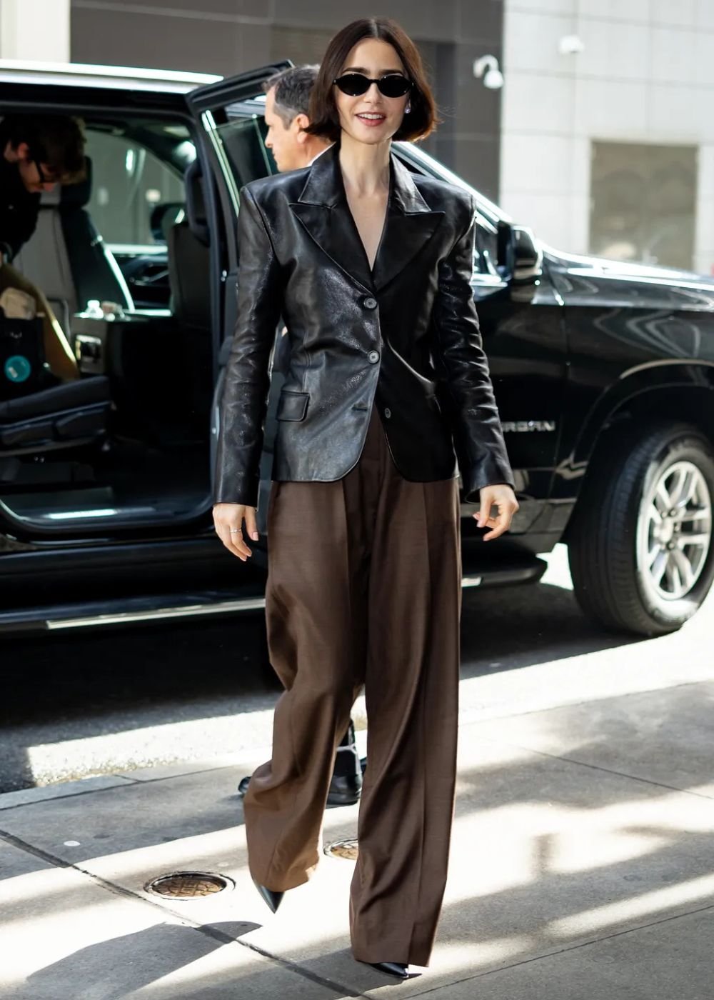 Mulher usando look com calça marrom larga e blazer de couro preto, caminhando na rua, próxima a um carro.