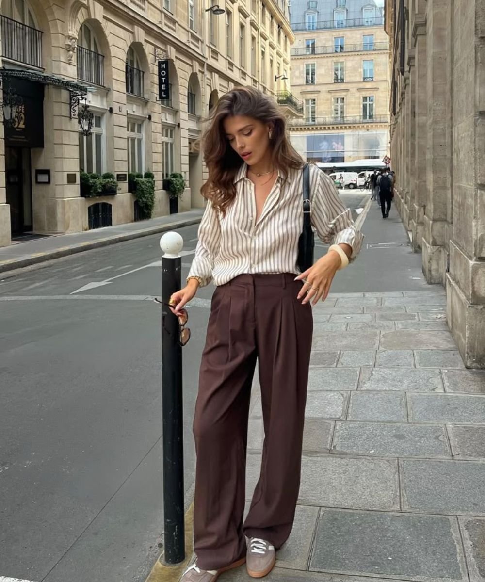 Pessoa em rua urbana, usando look com calça marrom larga, camisa listrada e tênis cinza.