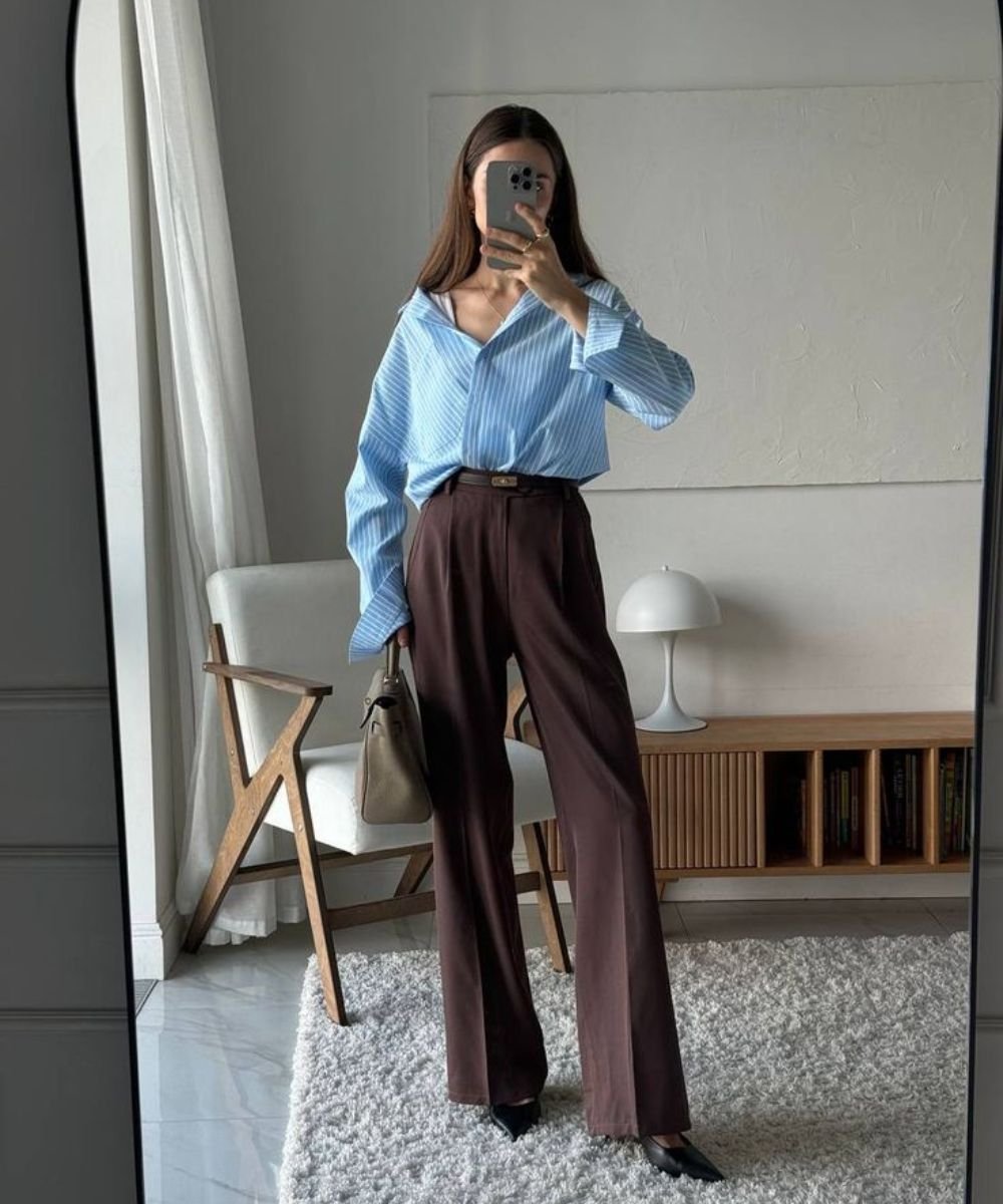 Pessoa vestindo look com calça marrom, blusa azul listrada e segurando uma bolsa, refletida em espelho.