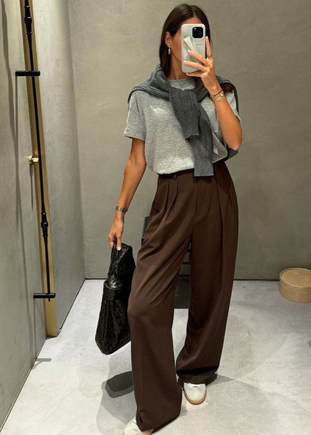Mulher com calça marrom e camiseta cinza, suéter nos ombros. Ela segura uma bolsa preta e usa tênis brancos.