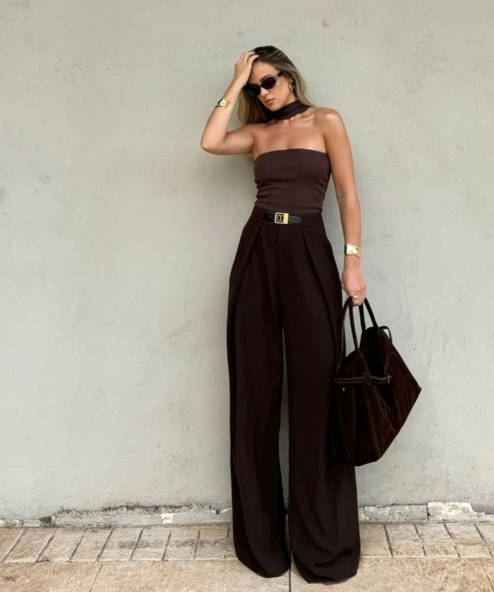 Pessoa de óculos de sol com top e calça marrom, segurando bolsa grande, em um look elegante.