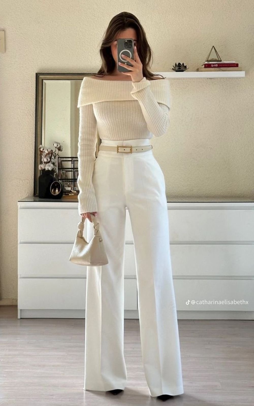 Modelo com look elegante em calça pantalona branca, blusa ombro a ombro e bolsa pequena, em frente a um espelho.