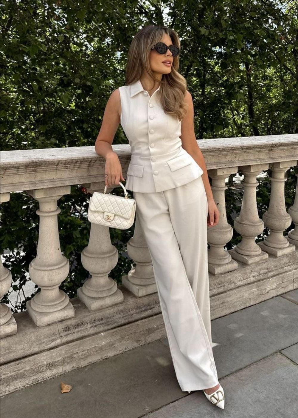 A pessoa está em uma varanda usando um look elegante com calça pantalona branca e colete, segurando uma bolsa.