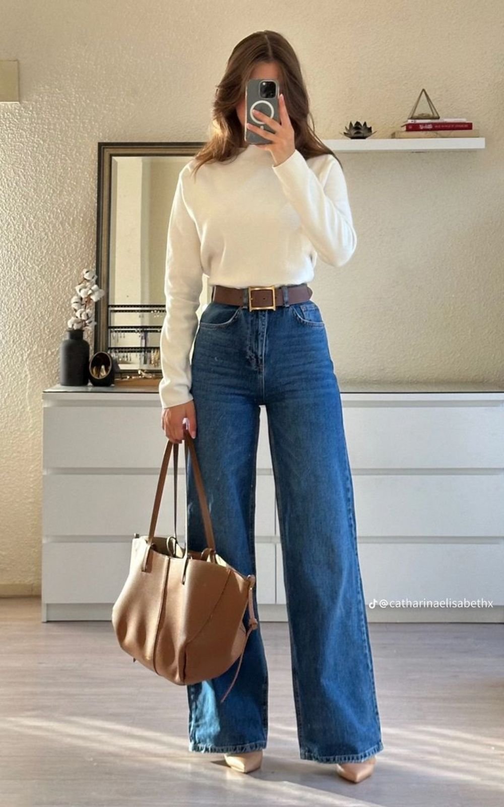 Pessoa veste look com calça pantalona jeans e blusa branca, segura bolsa marrom em ambiente interno.