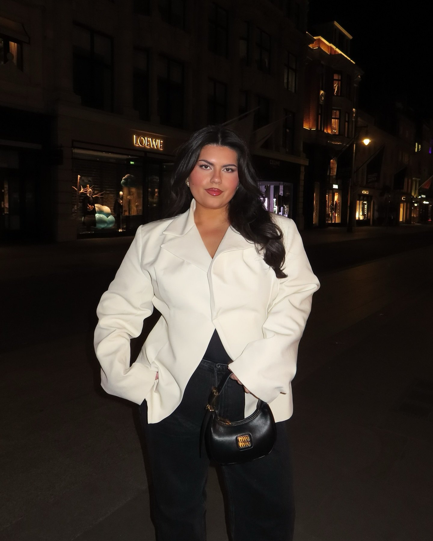 Pessoa em uma rua, à noite, vestindo blazer branco e segurando uma bolsa preta.