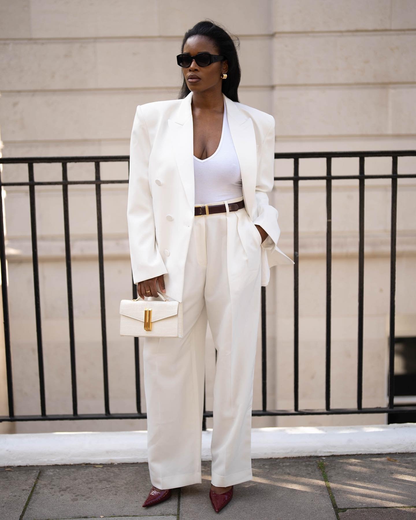 Pessoa usando blazer branco, blusa sem mangas e calça combinando, segurando bolsa elegante. Ela usa óculos escuros.