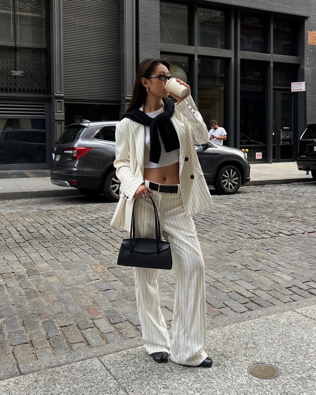 Pessoa com blazer branco listrado toma café na rua, carregando bolsa preta e usando óculos escuros.