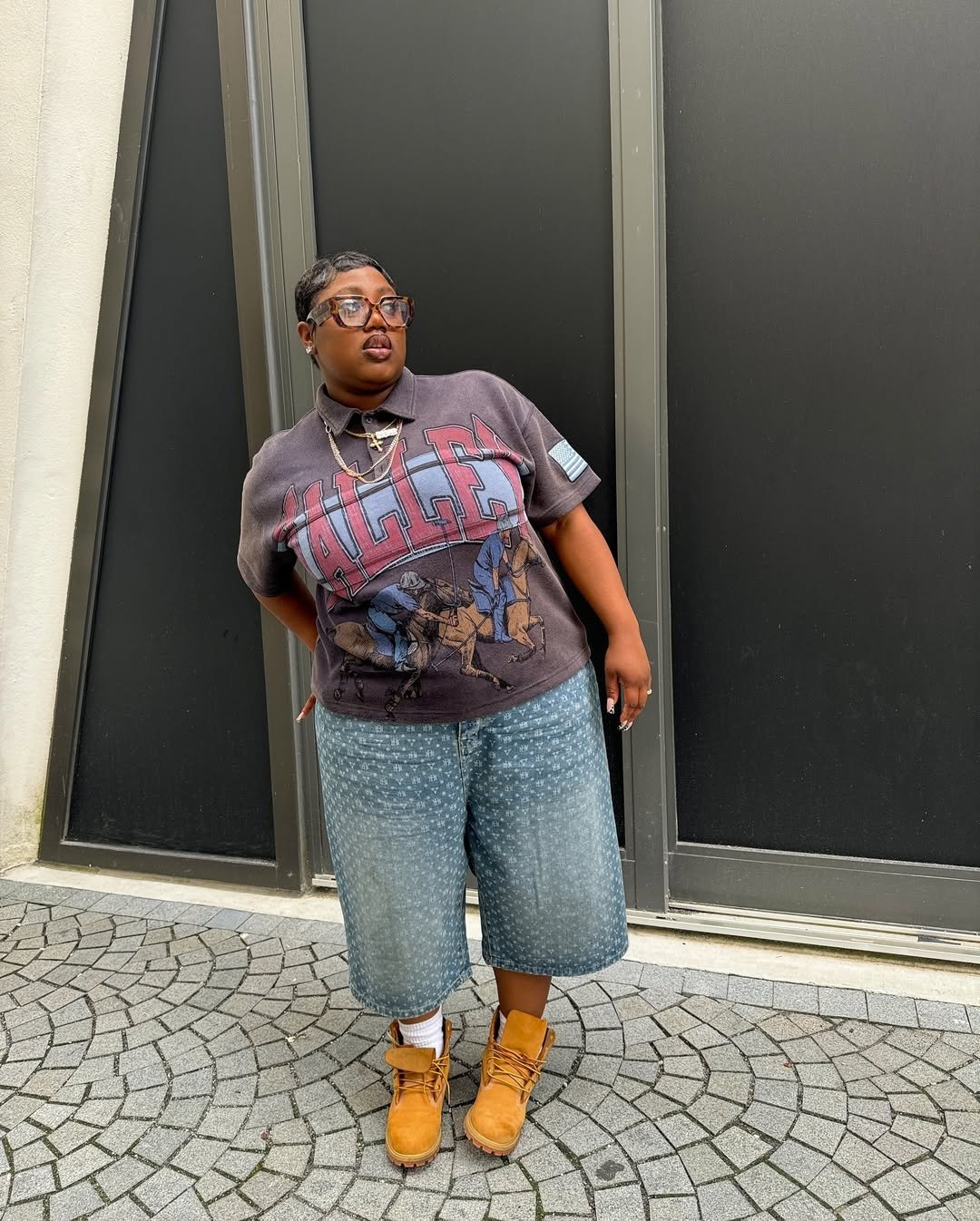 Pessoa vestindo camiseta estampada, bermuda jeans e bota Timberland, em pose descontraída diante de parede preta.