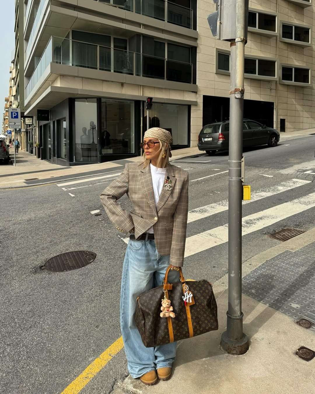 Pessoa em uma esquina de rua, usando jaqueta xadrez, calça jeans larga e carregando bolsa grande.