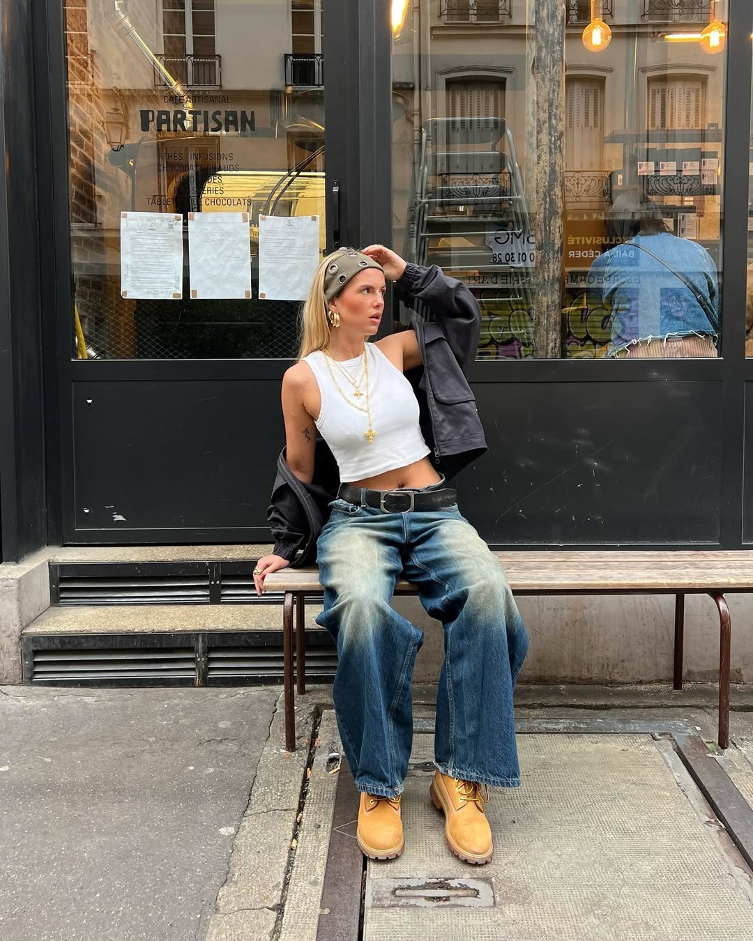 Pessoa sentada em banco, usando bota Timberland, calça jeans larga e top branco. Fundo com fachada de café.