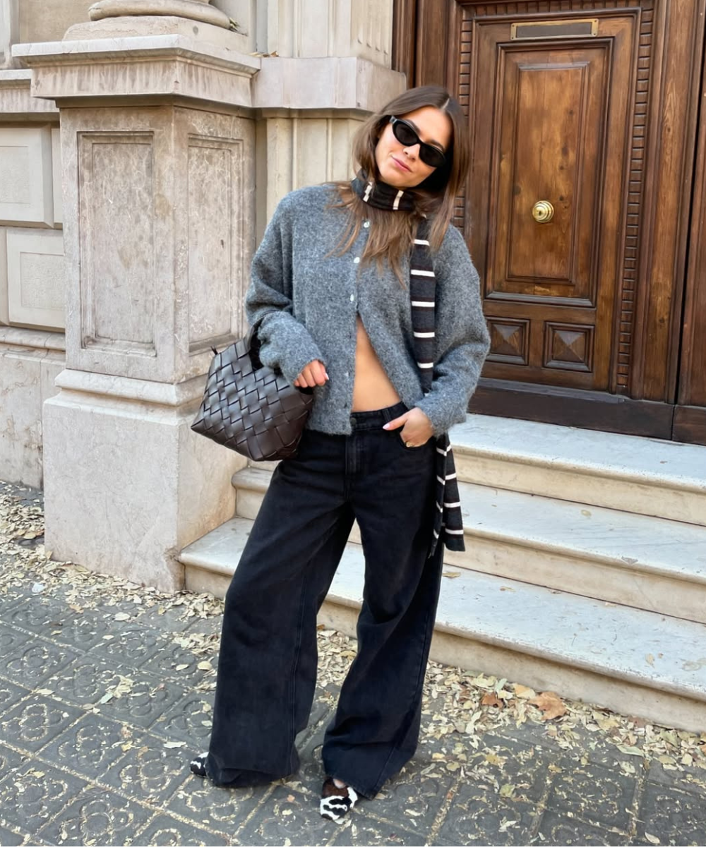 Pessoa de óculos escuros usa tricot cinza, calça larga preta, cachecol listrado e bolsa trançada em frente a porta de madeira. Exemplo de looks com cardigã.