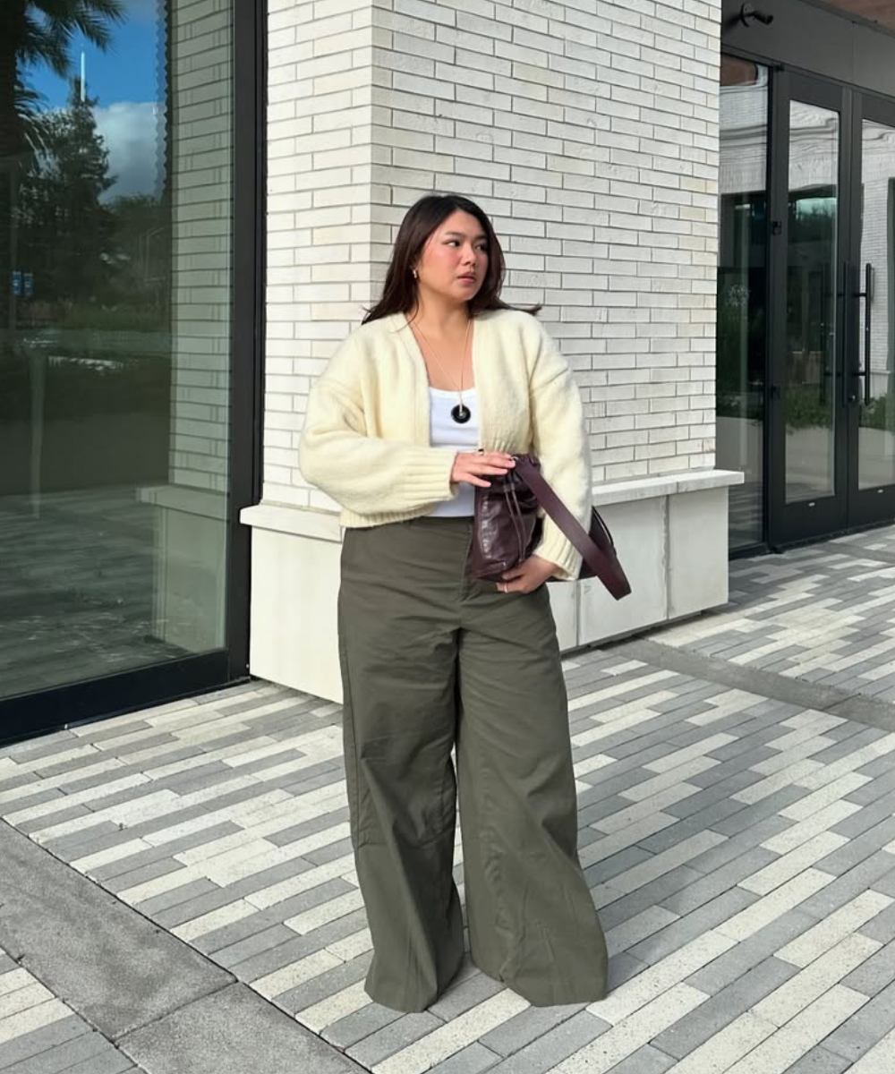 Pessoa com cardigã creme, segurando bolsa marrom, blusa branca e calça verde larga, em frente a um prédio de tijolos.