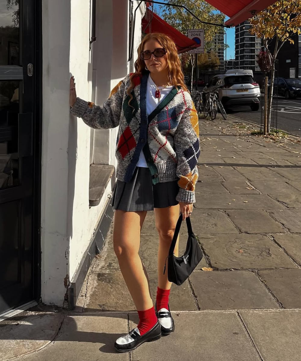 Pessoa usando cardigã xadrez colorido, saia cinza e meias vermelhas, em uma calçada urbana ensolarada.