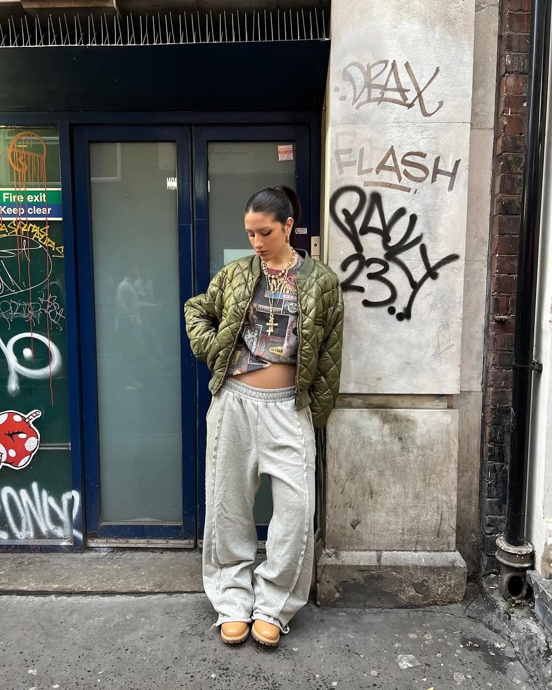 Pessoa com jaqueta acolchoada verde, camiseta estampada e calça larga cinza. Coturnos femininos bege. Graffiti ao fundo.
