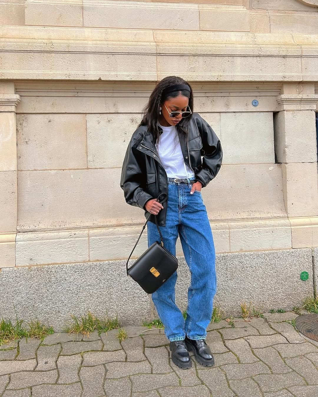 Mulher vestindo jaqueta preta, camiseta branca, jeans e coturno, com bolsa tiracolo. Looks urbanos e despojados.