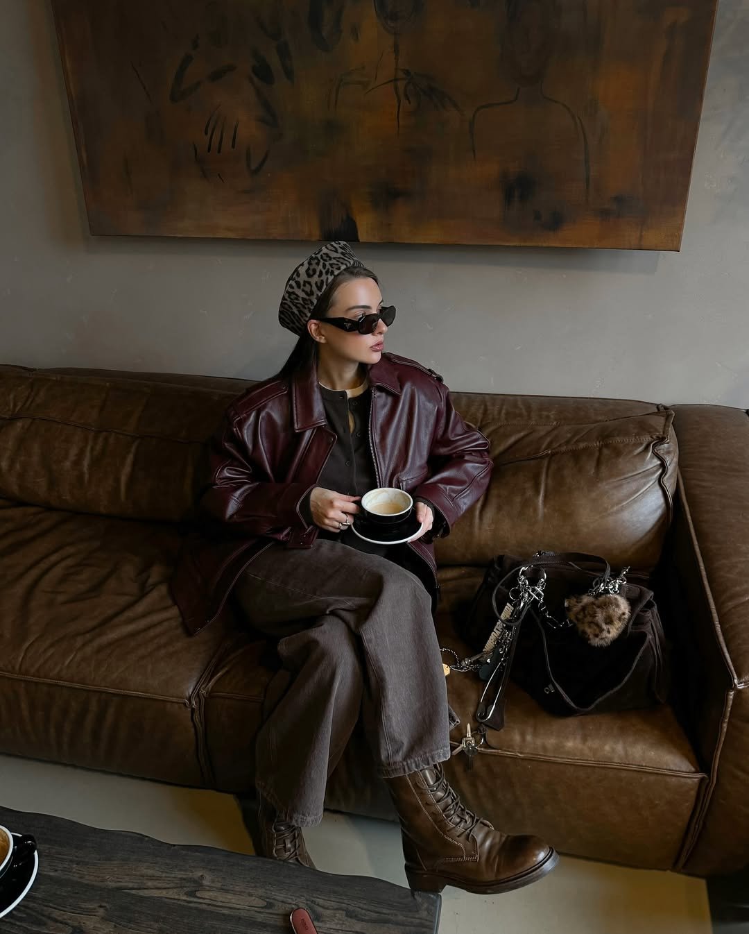 Pessoa em sofá com casaco marrom, boina de oncinha e coturno, segurando café. Look elegante e casual.