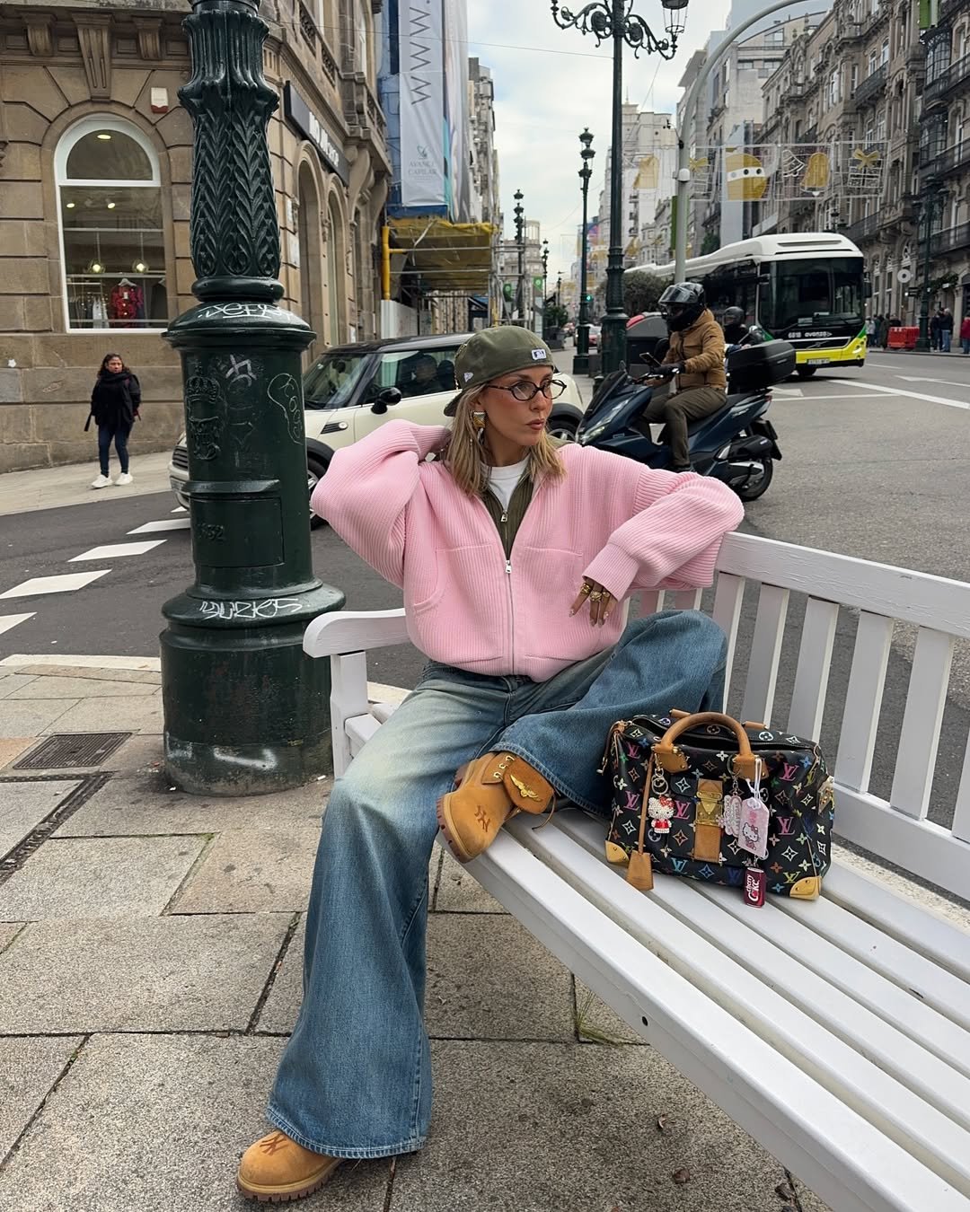 Pessoa com jaqueta rosa e jeans sentada em banco. Bolsa estilosa ao lado.