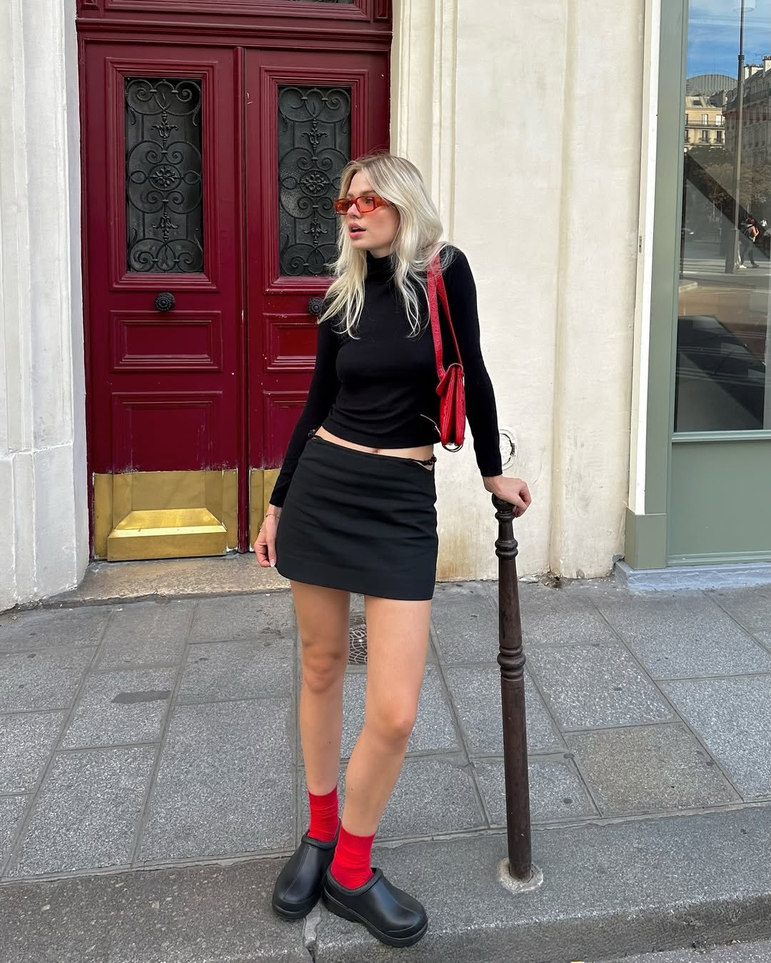 Pessoa usando meias vermelhas, sapatos pretos, saia e blusa pretas, óculos vermelhos e bolsa, em frente a uma porta vermelha.