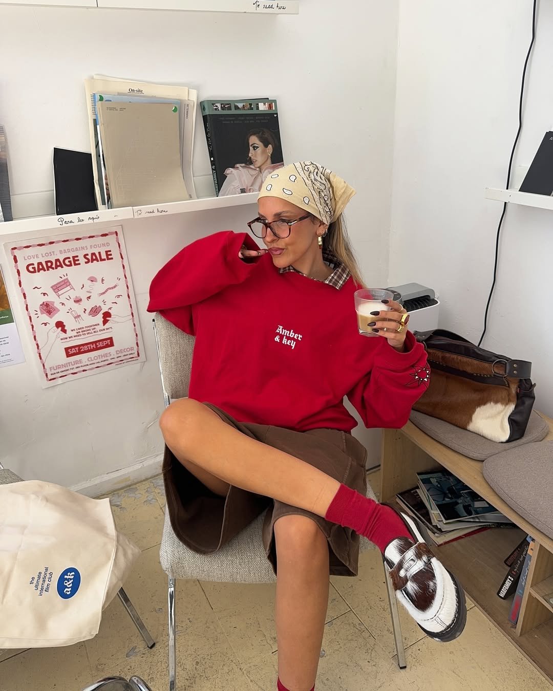 Pessoa sentada em cadeira, usando blusa vermelha, saia marrom e meia vermelha, segurando copo, ao lado de livros e bolsas.