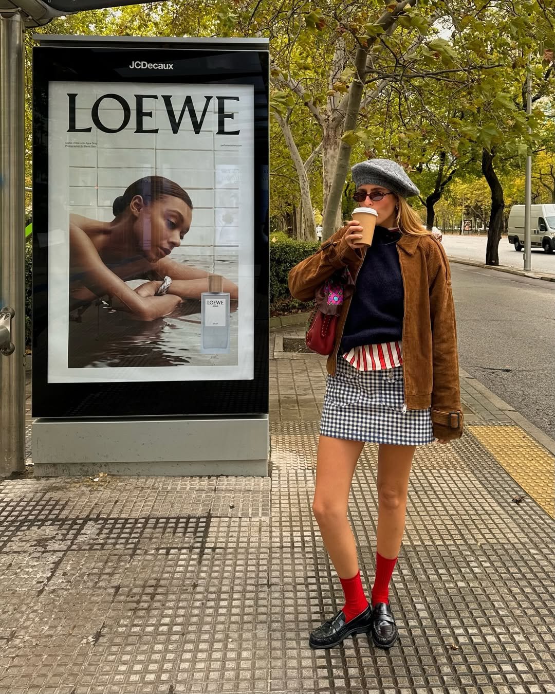 Uma pessoa com boina cinza, jaqueta marrom e meias vermelhas bebe café ao lado de um ponto de ônibus, com cartaz da Loewe.
