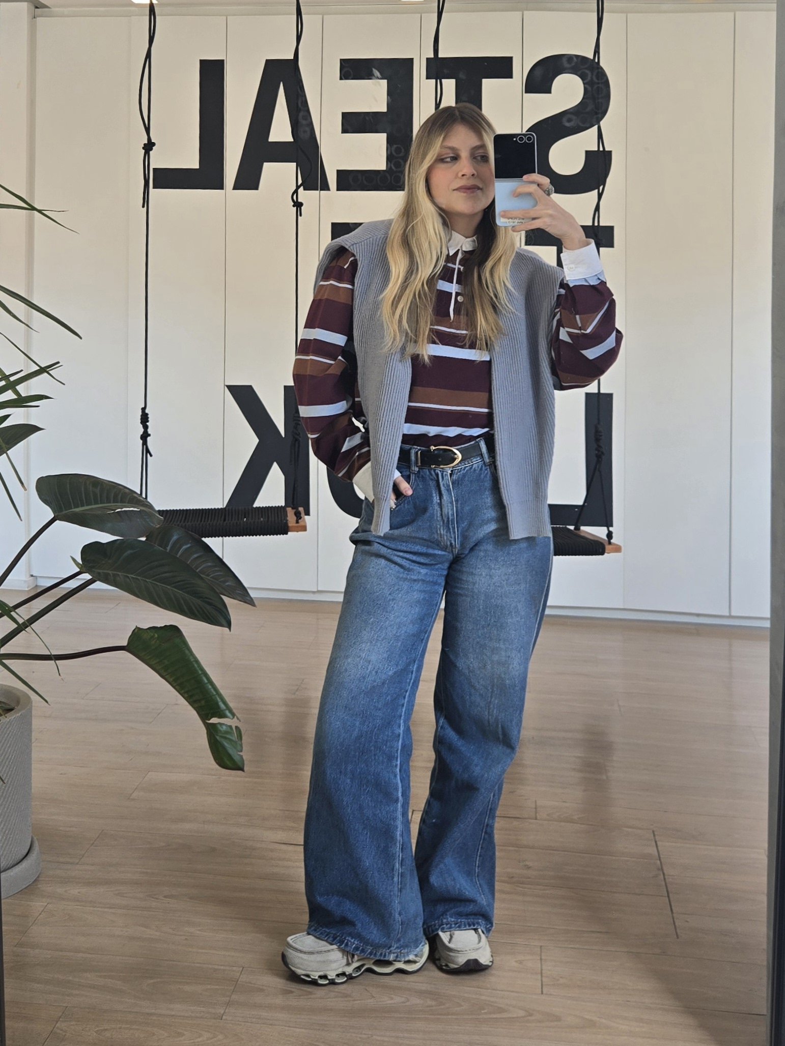 Pessoa vestida com blusa listrada e calça jeans, tirando selfie em um espaço moderno.