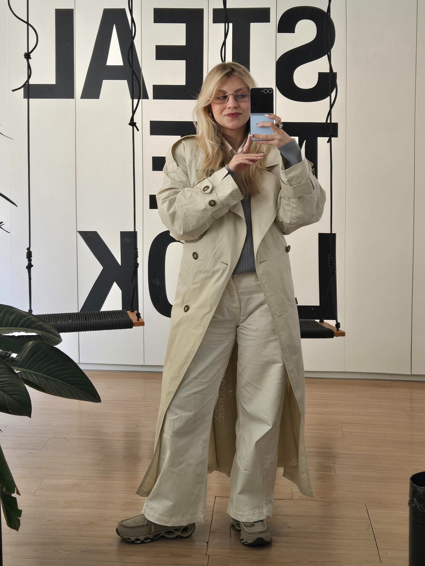 Mulher de casaco bege, calça branca e óculos, tirando selfie. Mocassins cinza completam o look tendência.